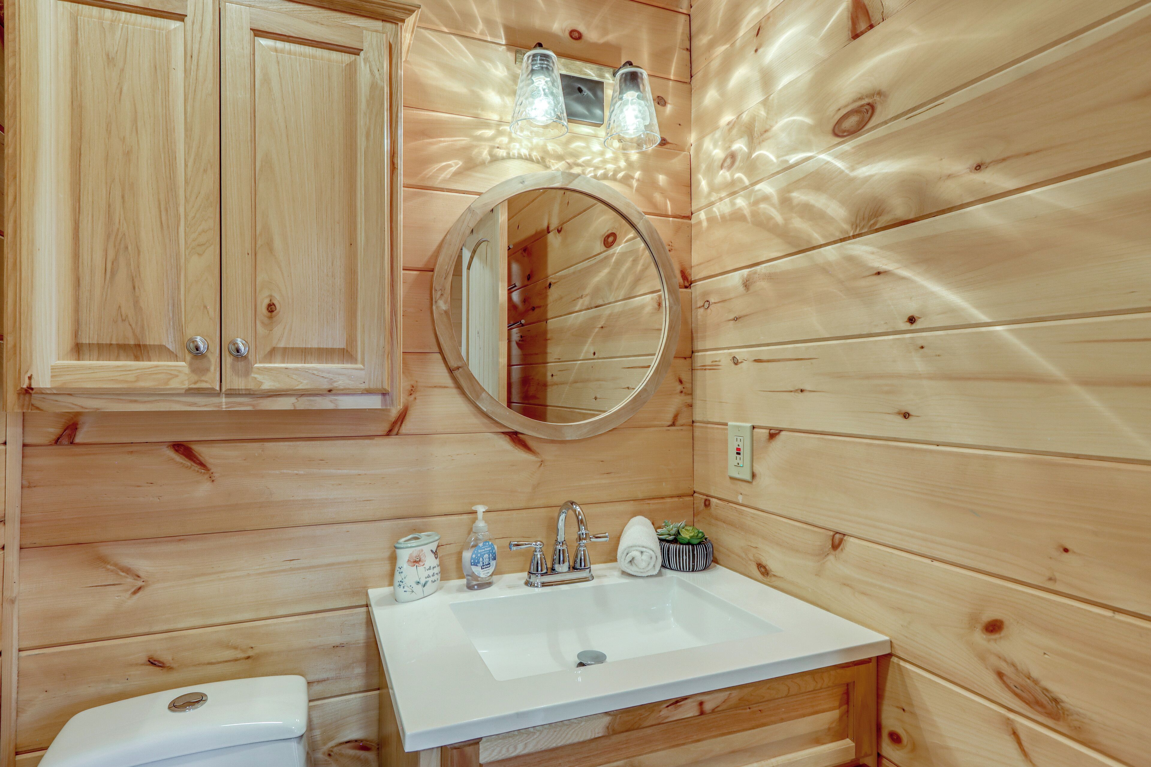 A wooden cabin bathroom with a white sink, two cabinets, a round mirror, and a small window with light fixtures.