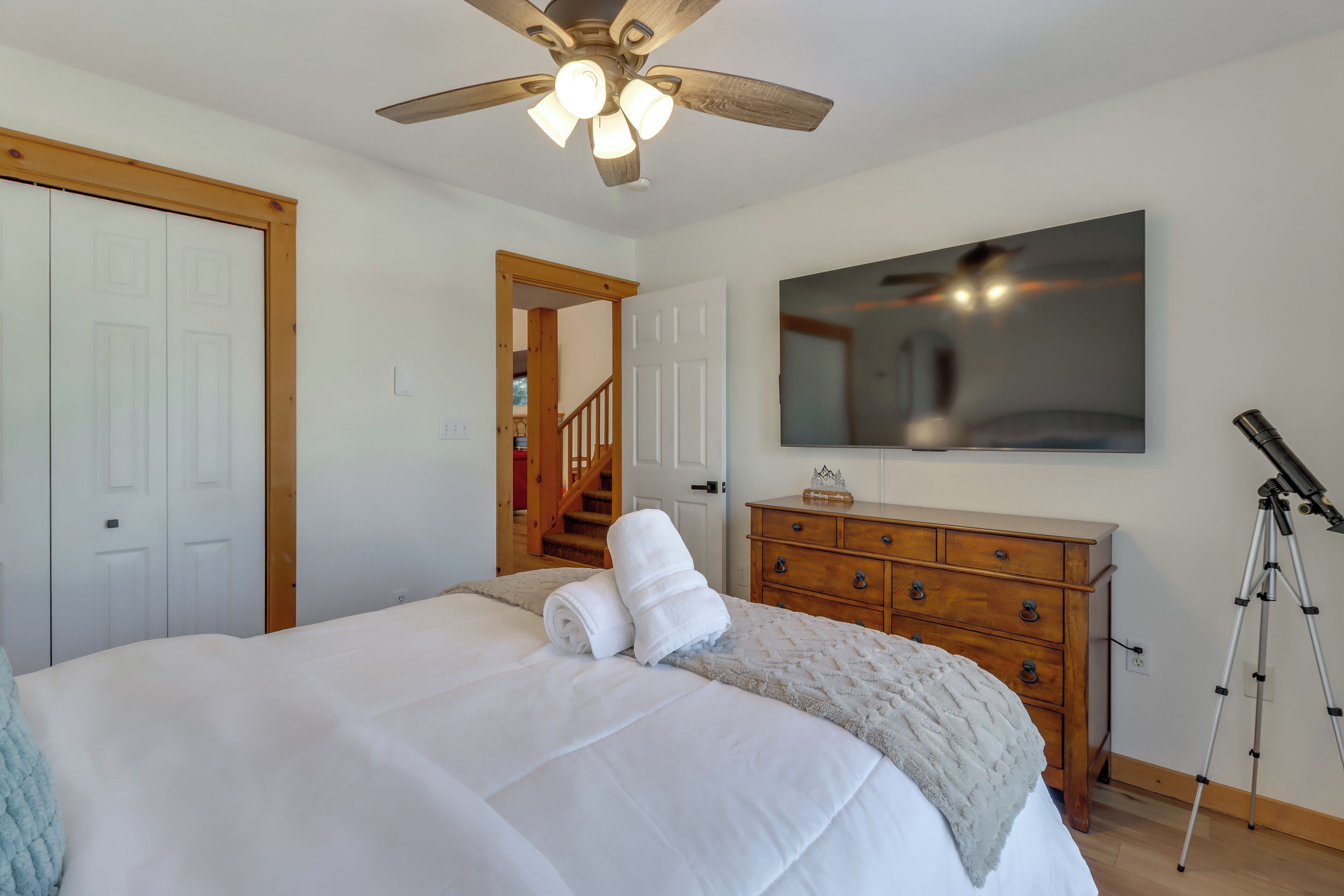 A bedroom with a bed, a ceiling fan, a dresser, a mirror, a telescope, and a staircase.