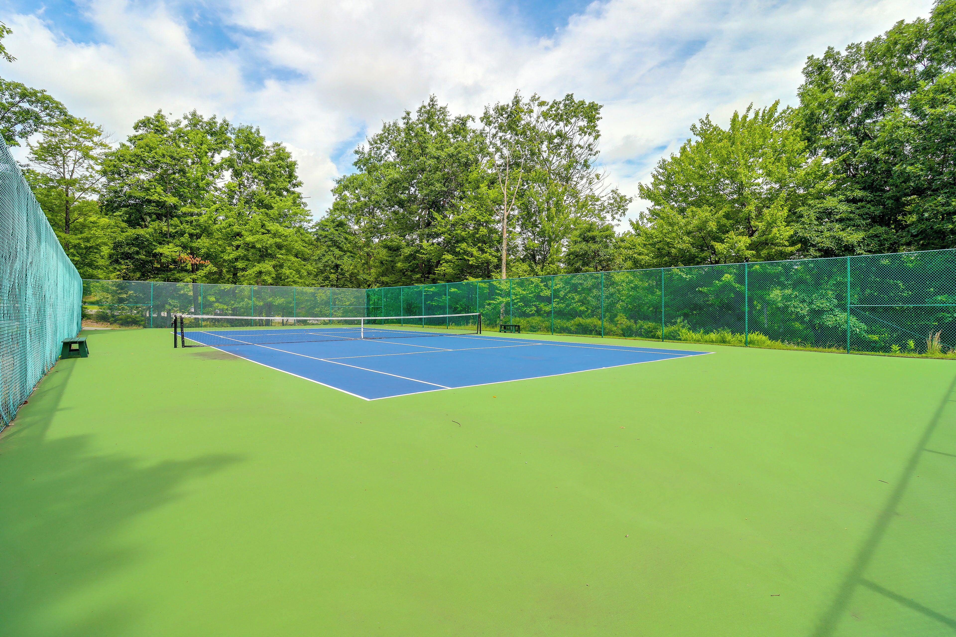 Ein Tennisplatz mit Netz, umgeben von grünen Zäunen und Bäumen.