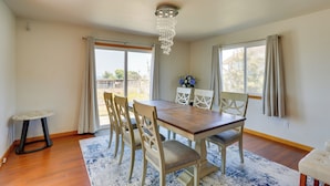 A dining area with a wooden table, six chairs, a chandelier, and large windows with curtains.