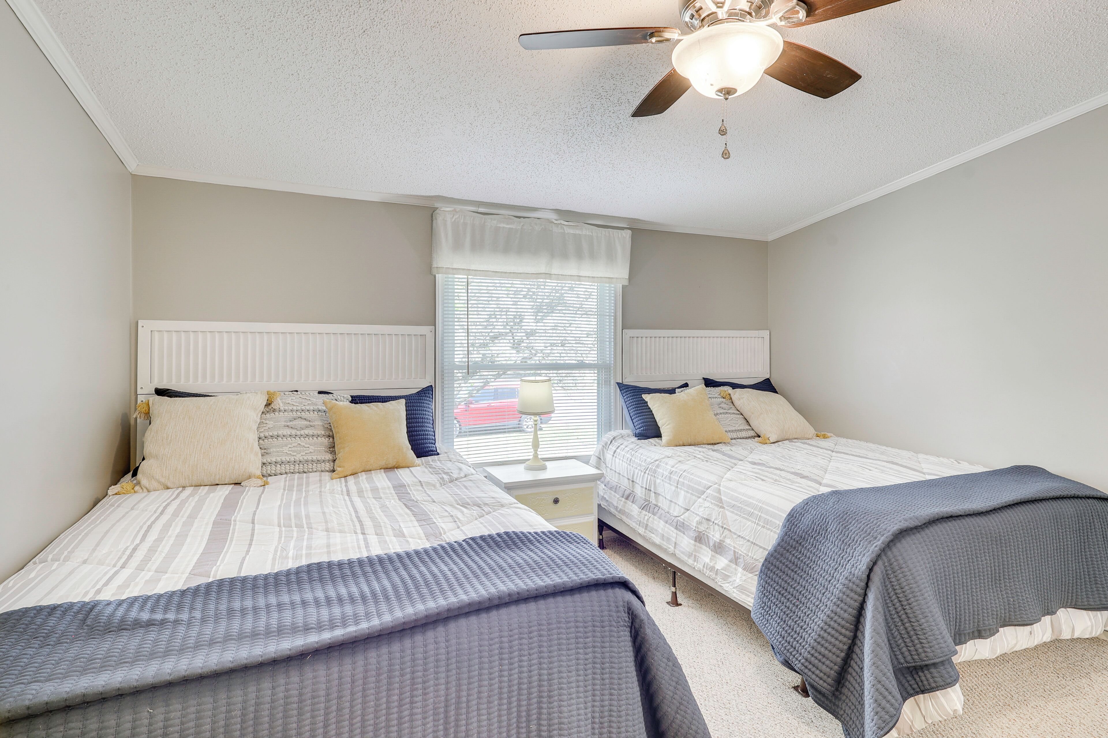 A bedroom with two beds, a ceiling fan, a window with blinds, and a nightstand with a lamp.