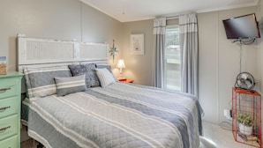 A bedroom with a bed, a ceiling fan, a TV, a nightstand, a dresser, and a window with curtains. - Nina's Beach Retreat: ~ 1 Mi to Holden Beach! (Supply)