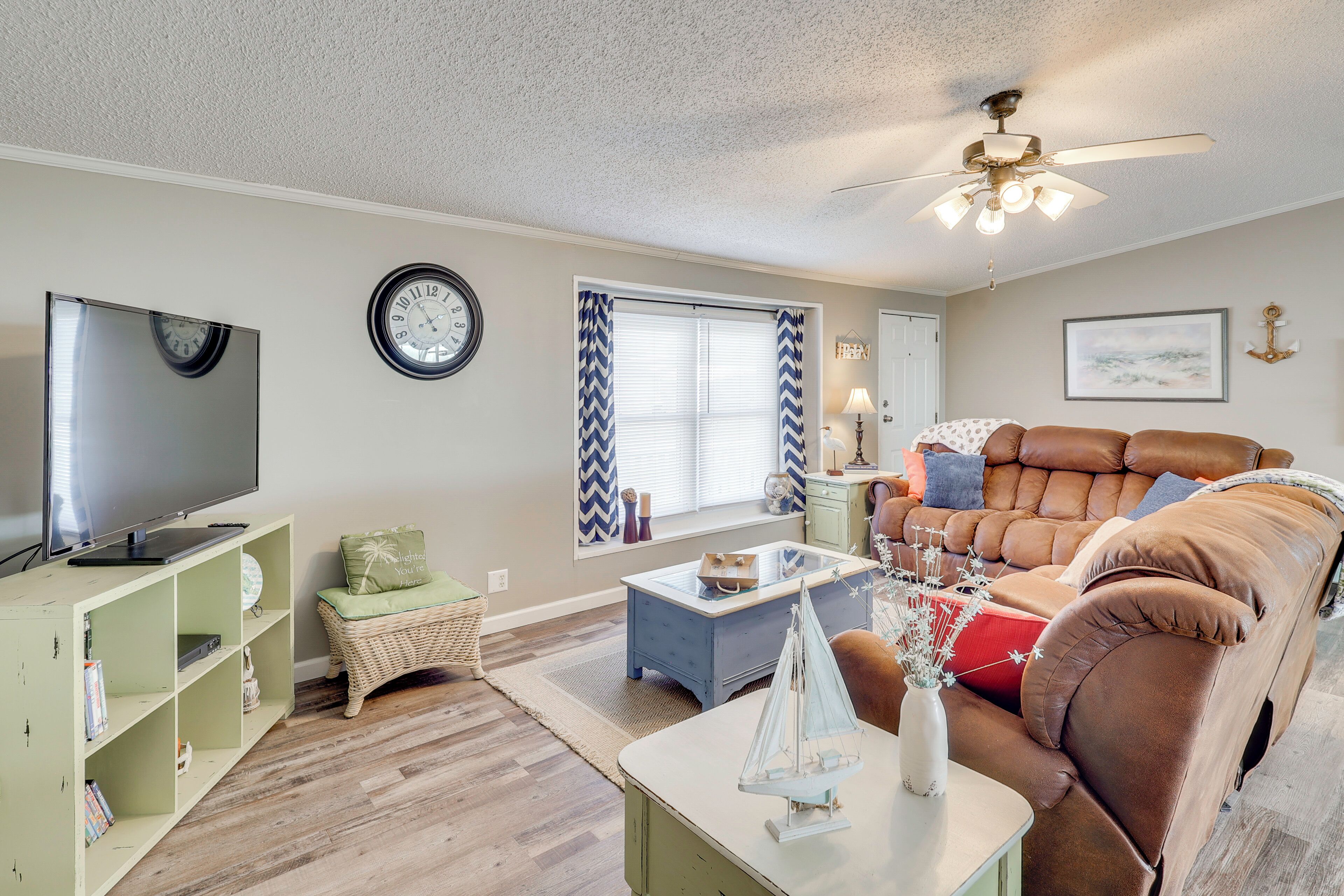 A living room with a flat-screen TV, a sofa, a coffee table, a small table with a decorative item, a wall clock, and a window with curtains.