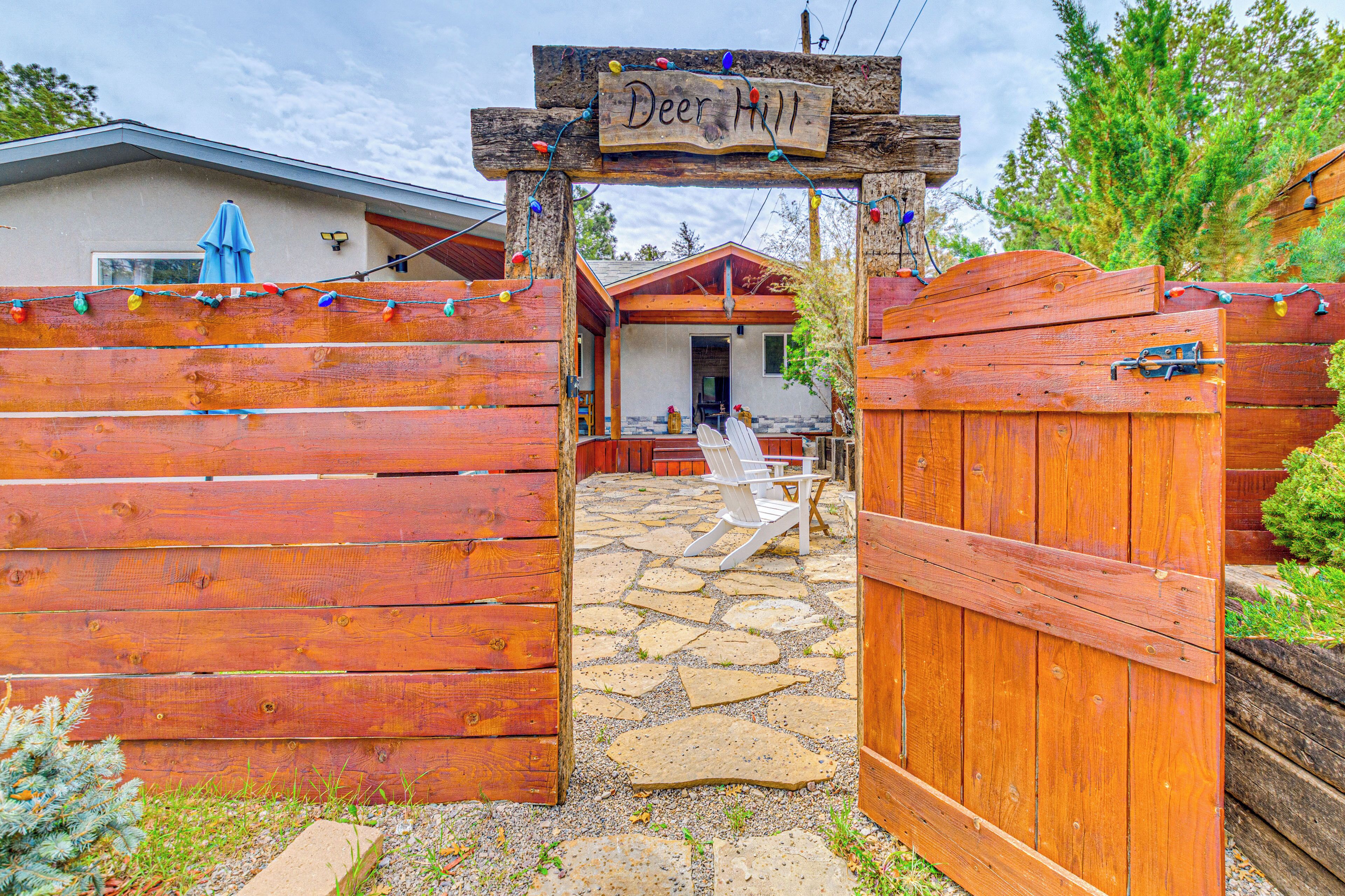 A rustic wooden gate with a sign reading 'Deer Hill,' leading to a house with a stone patio and a white chair outside.