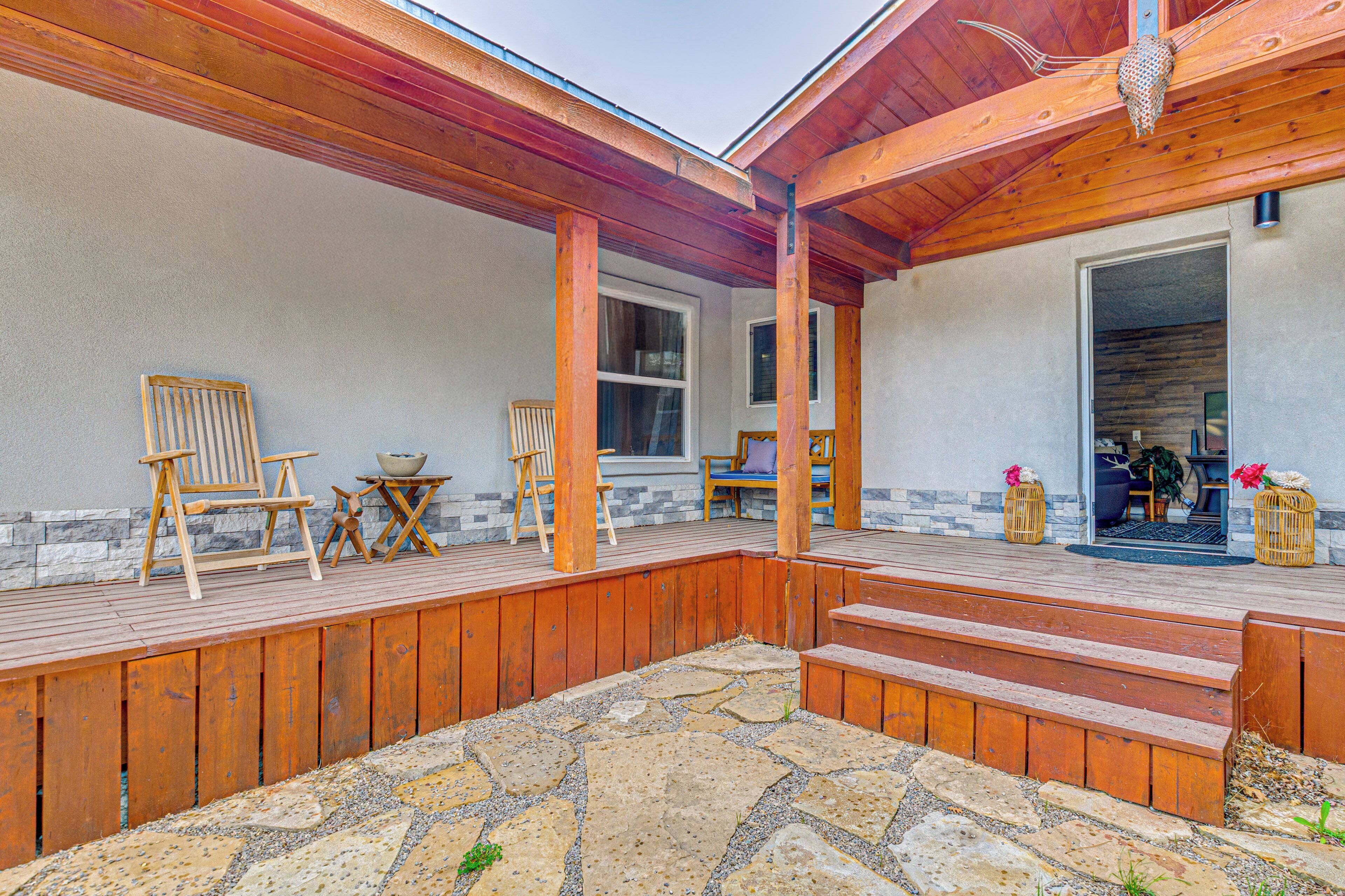 A covered outdoor deck with wooden furniture, stone flooring, and a view into the house.