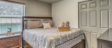 A bedroom with a bed, wooden nightstand, dresser, and a window with blinds.