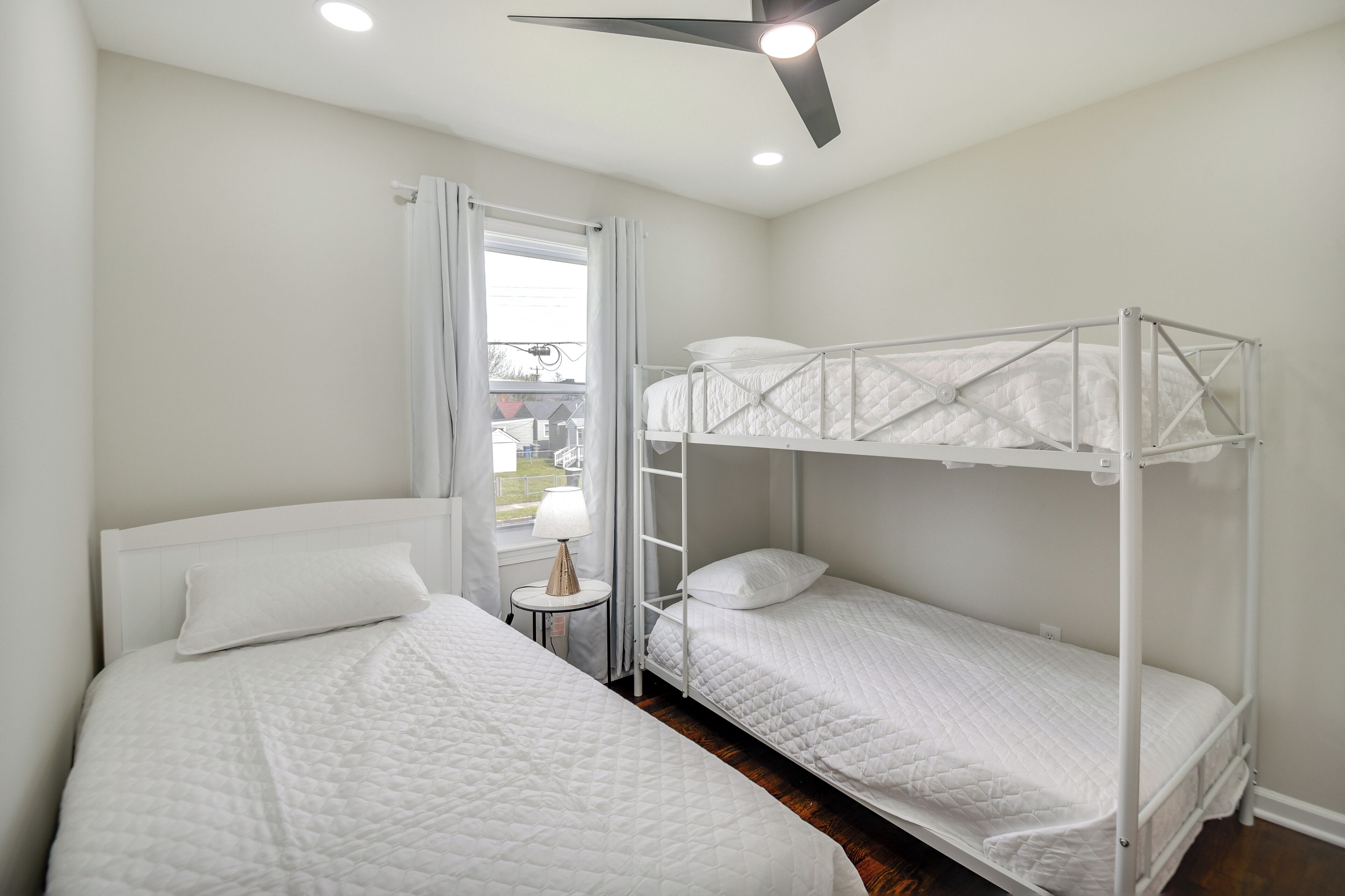 A room with two beds, one with a headboard and the other with a bunk bed, a ceiling fan, and a window with curtains.