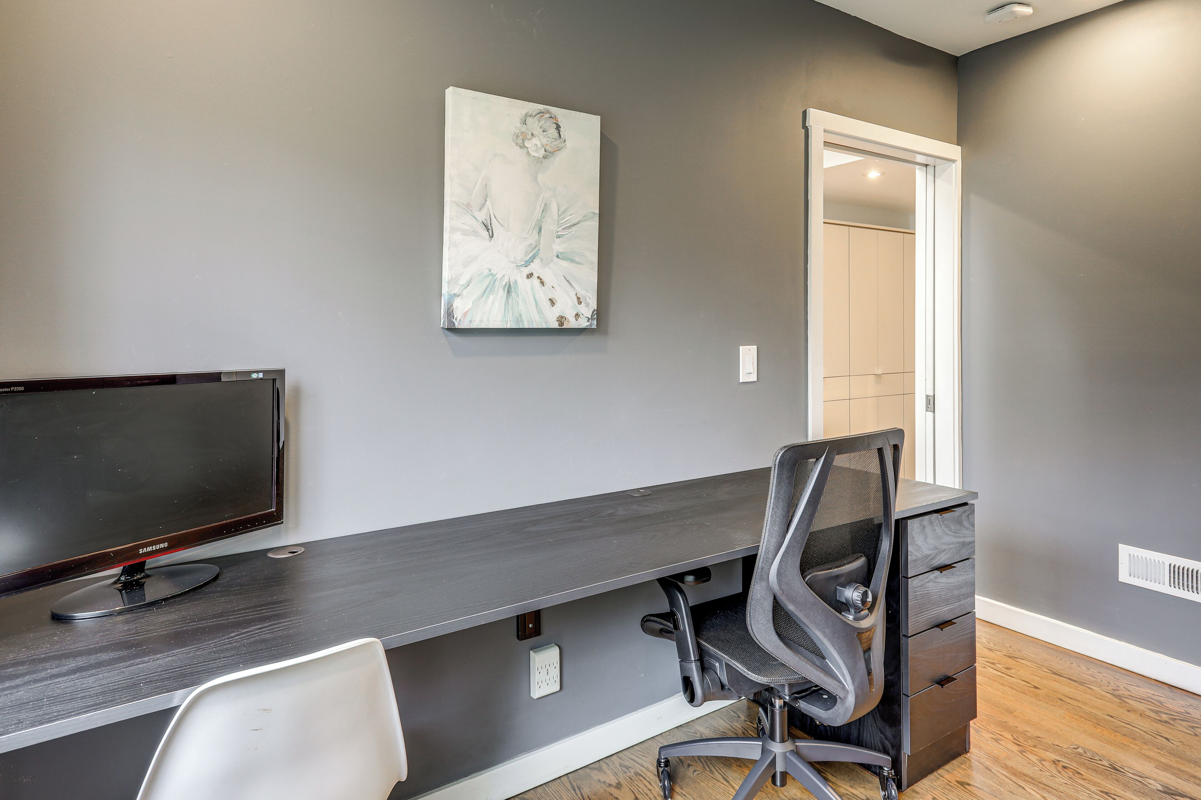 A modern office space with a long desk, a computer monitor, a chair, and a painting on the wall.