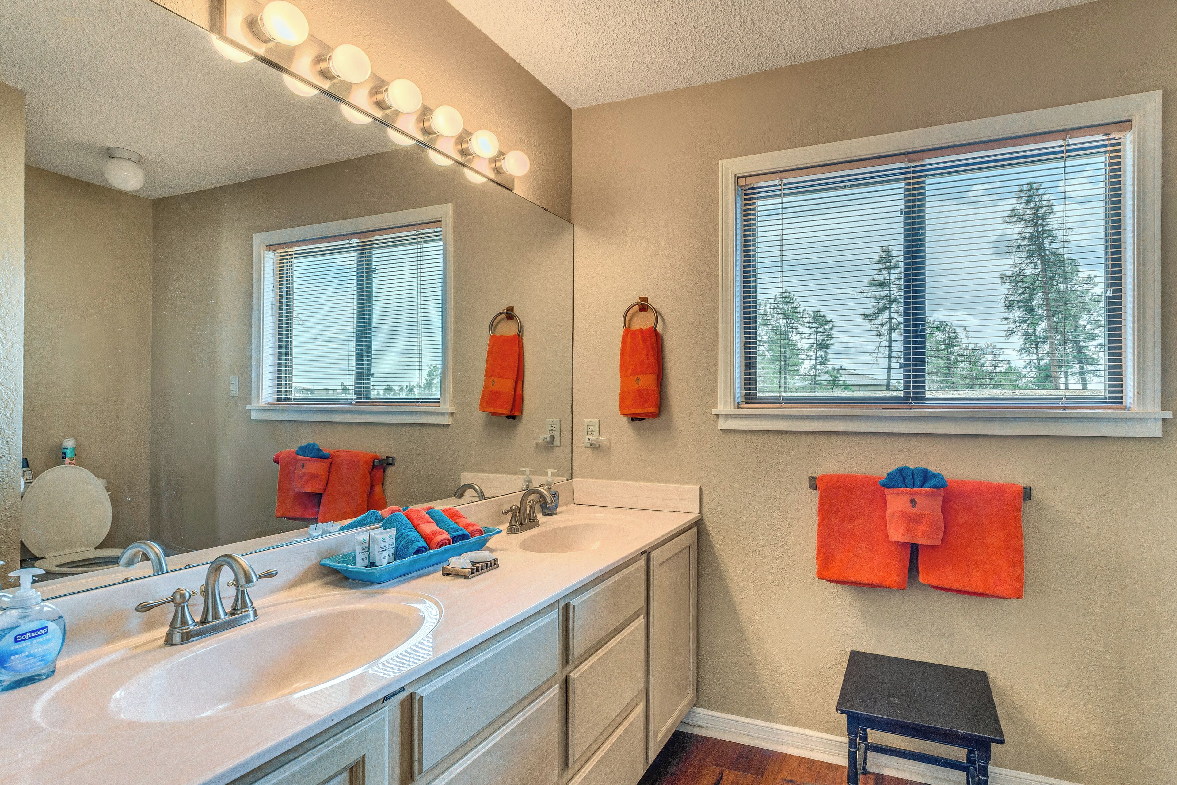 A bathroom with a double sink, vanity, mirror with lights, towel racks, and a window with blinds.