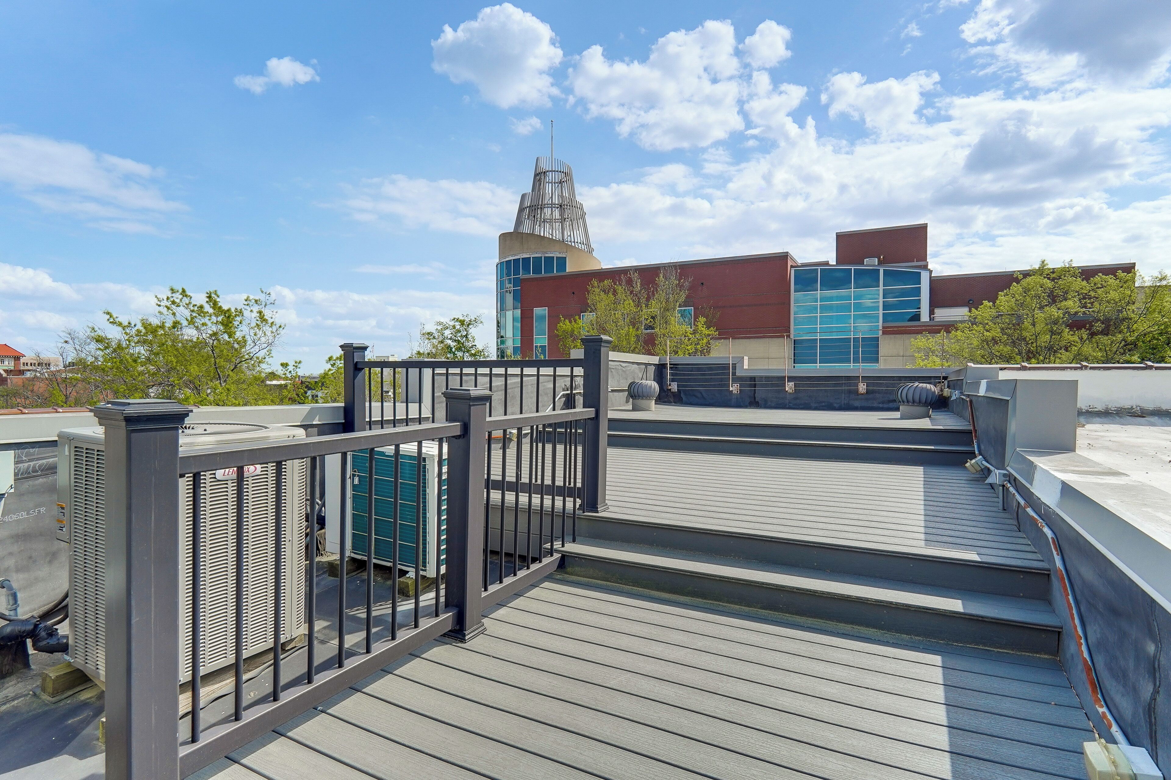 A rooftop deck with a metal railing, HVAC units, and a view of buildings in the background.