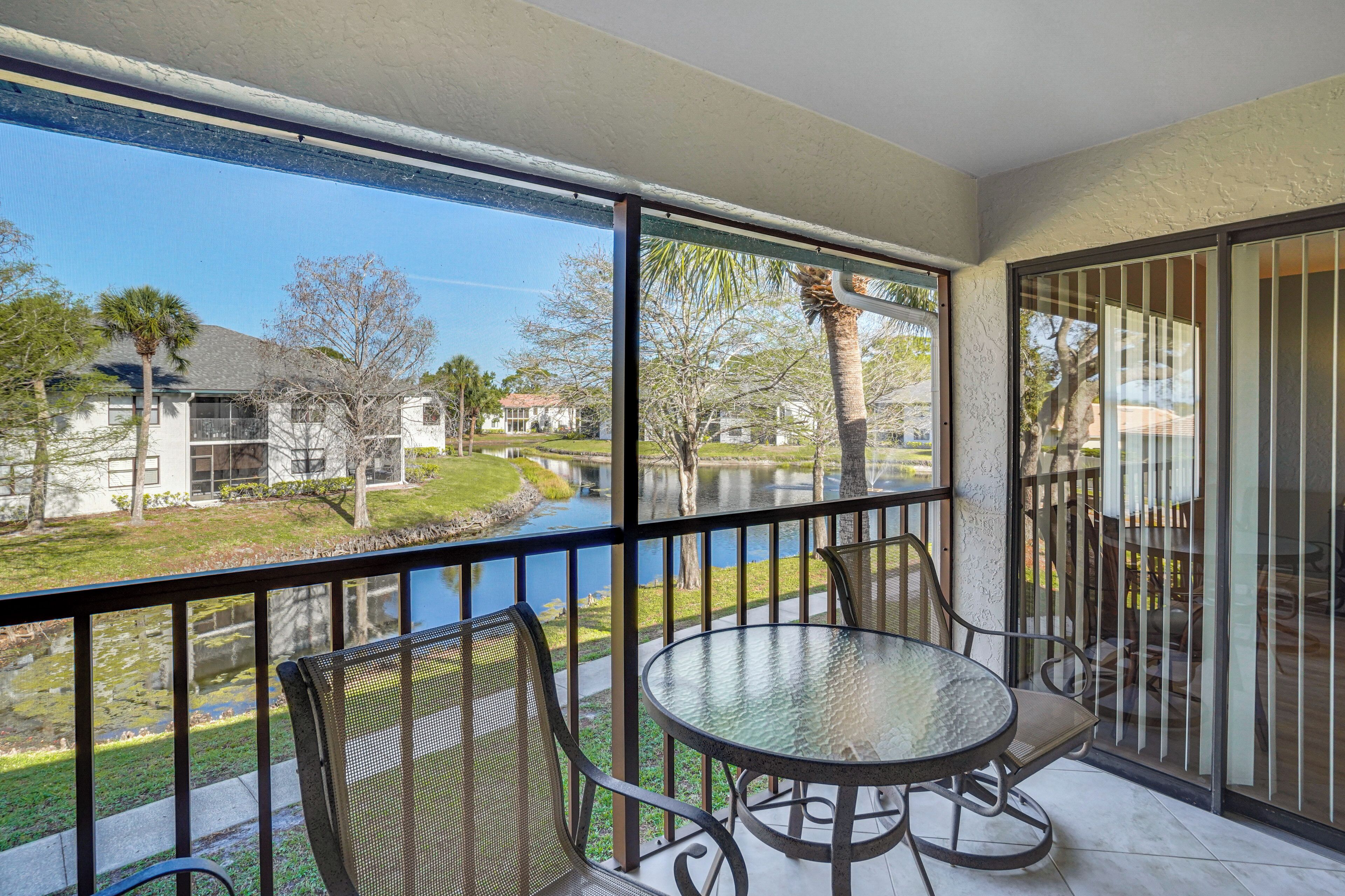 A balcony with a glass table and chairs, overlooking a water body and residential buildings.