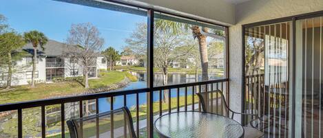 A balcony with a glass table and chairs, overlooking a water body and residential buildings.