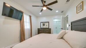 Une chambre à coucher comprenant un lit, un ventilateur de plafond, une télévision, une commode et une porte donnant sur une salle de bain.