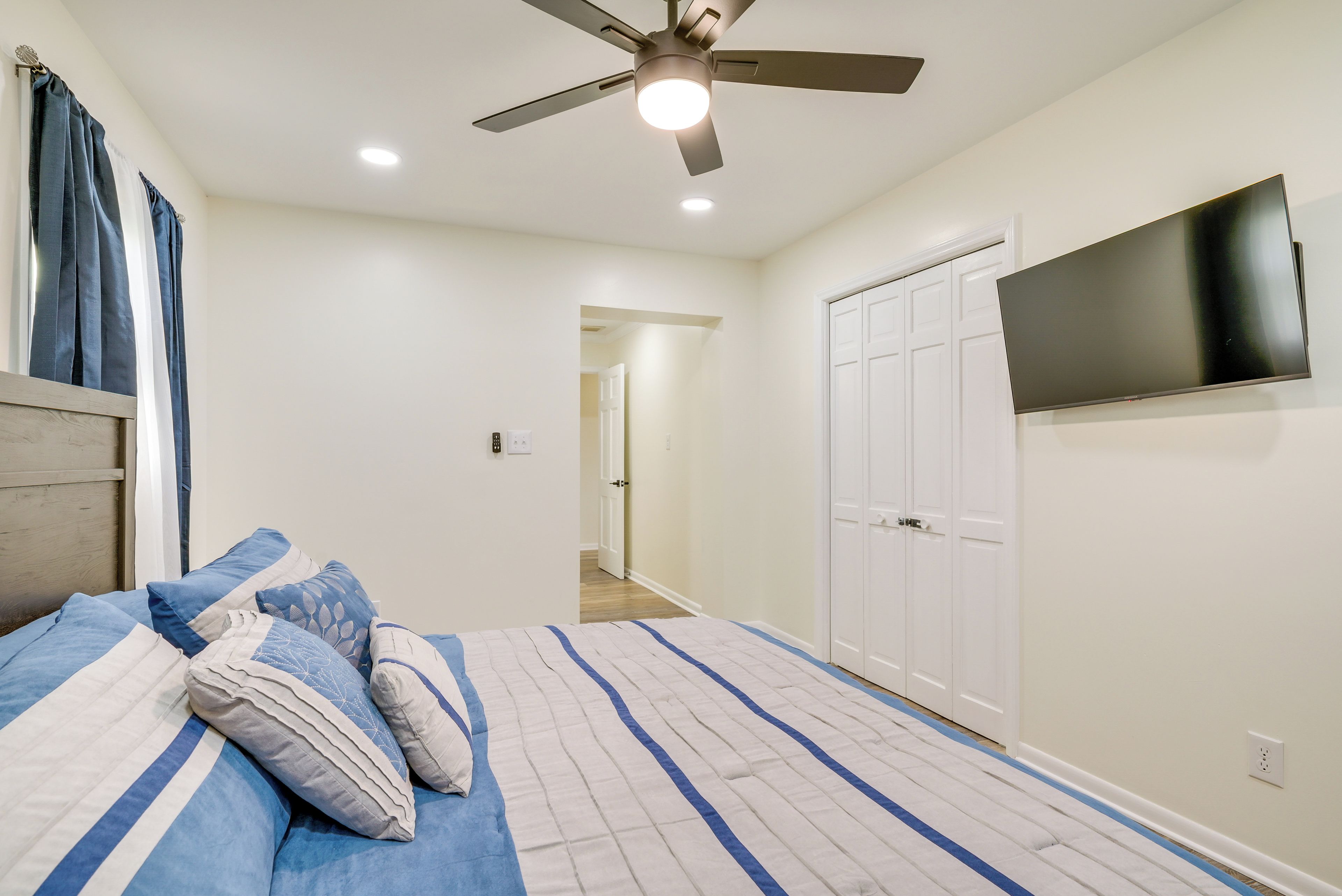 A bedroom with a bed, ceiling fan, TV, wardrobe, and a hallway leading to another room.