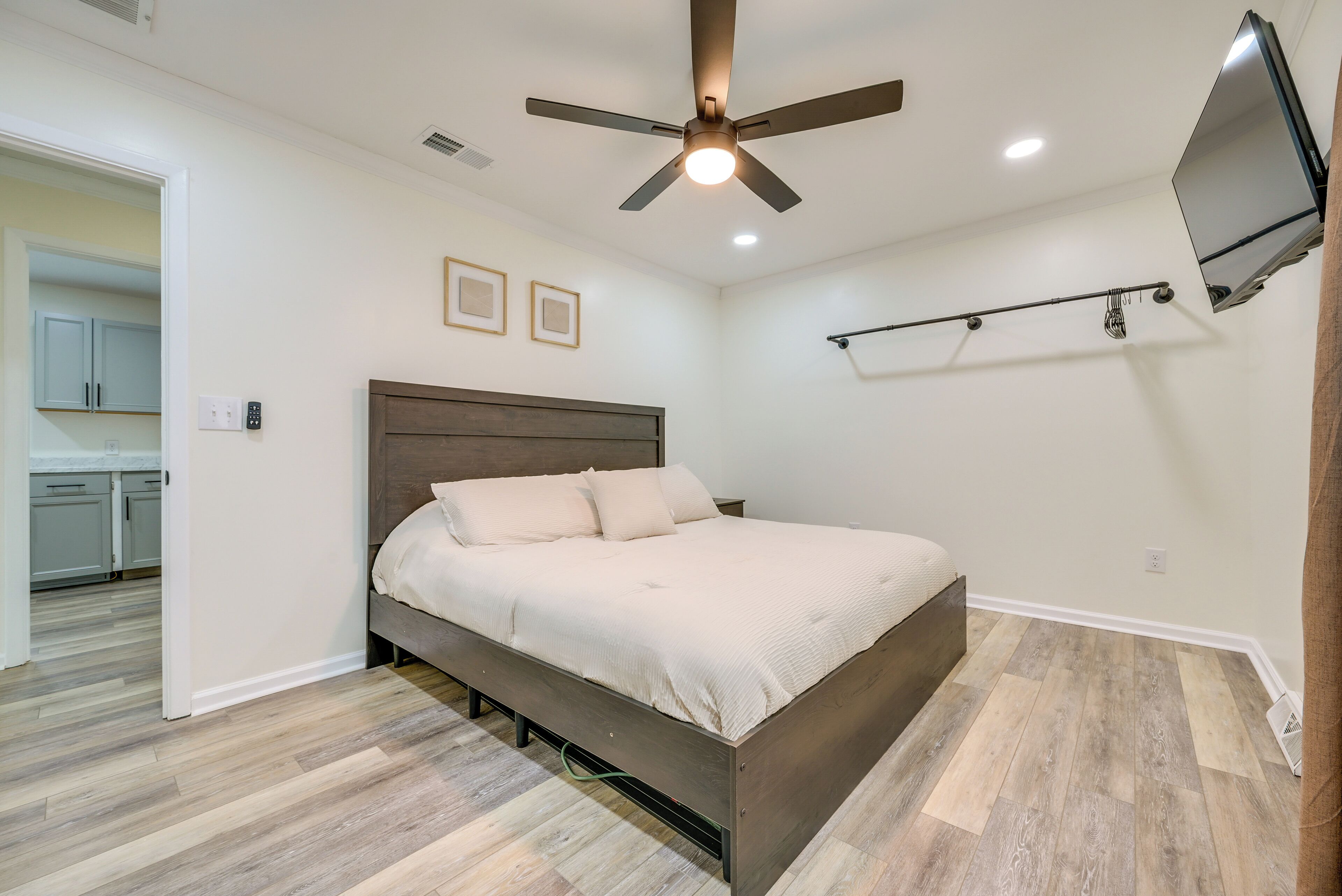 Quarto com cama, ventilador de teto, televisão na parede e cozinha visível através de uma porta aberta.