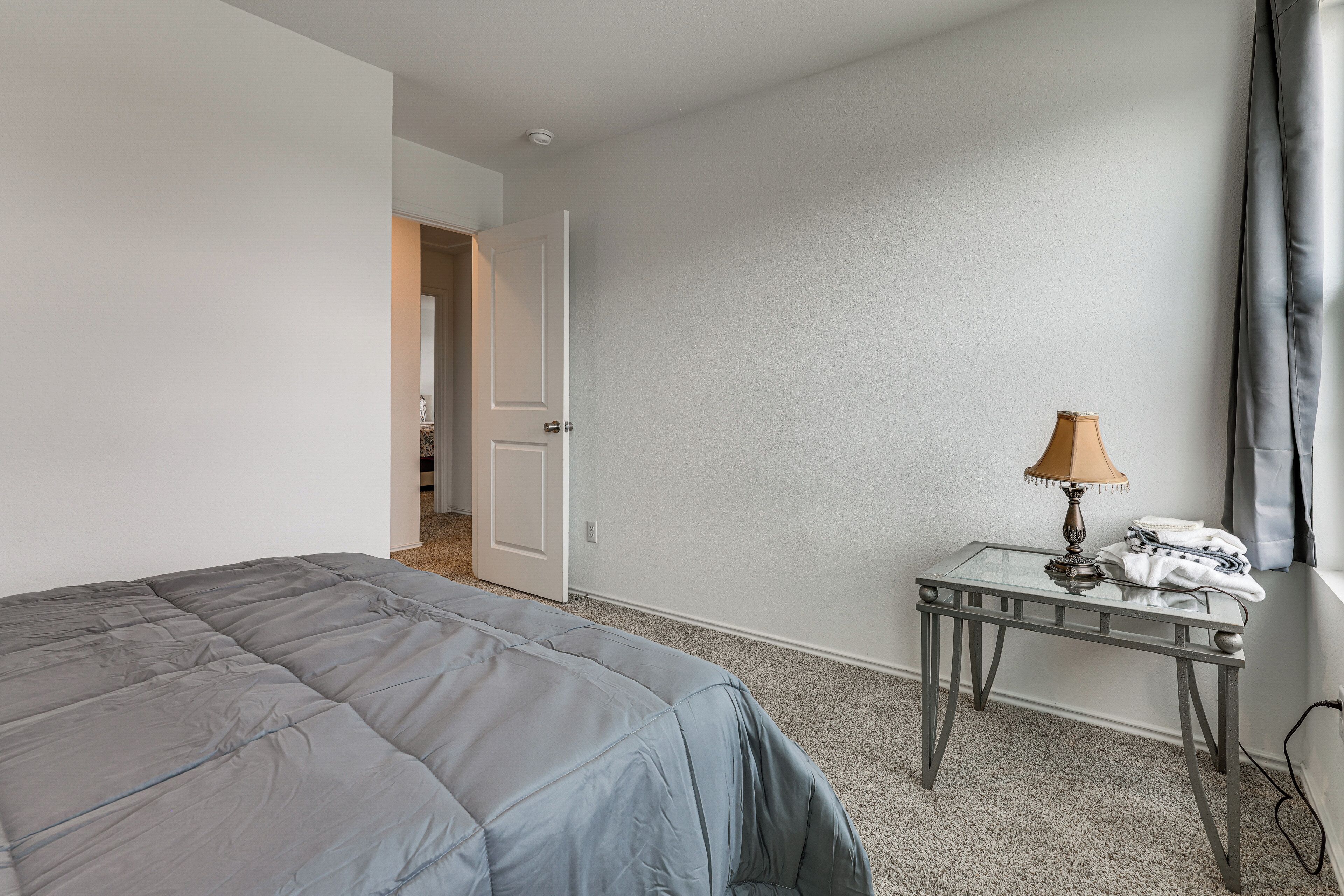 Um quarto com uma cama, uma mesa com abajur e uma porta que dá para outro cômodo.