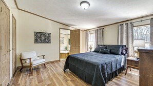 Une chambre avec un plancher en bois, un lit recouvert d’une couverture foncée, une chaise blanche, une armoire en bois et une fenêtre avec des rideaux.