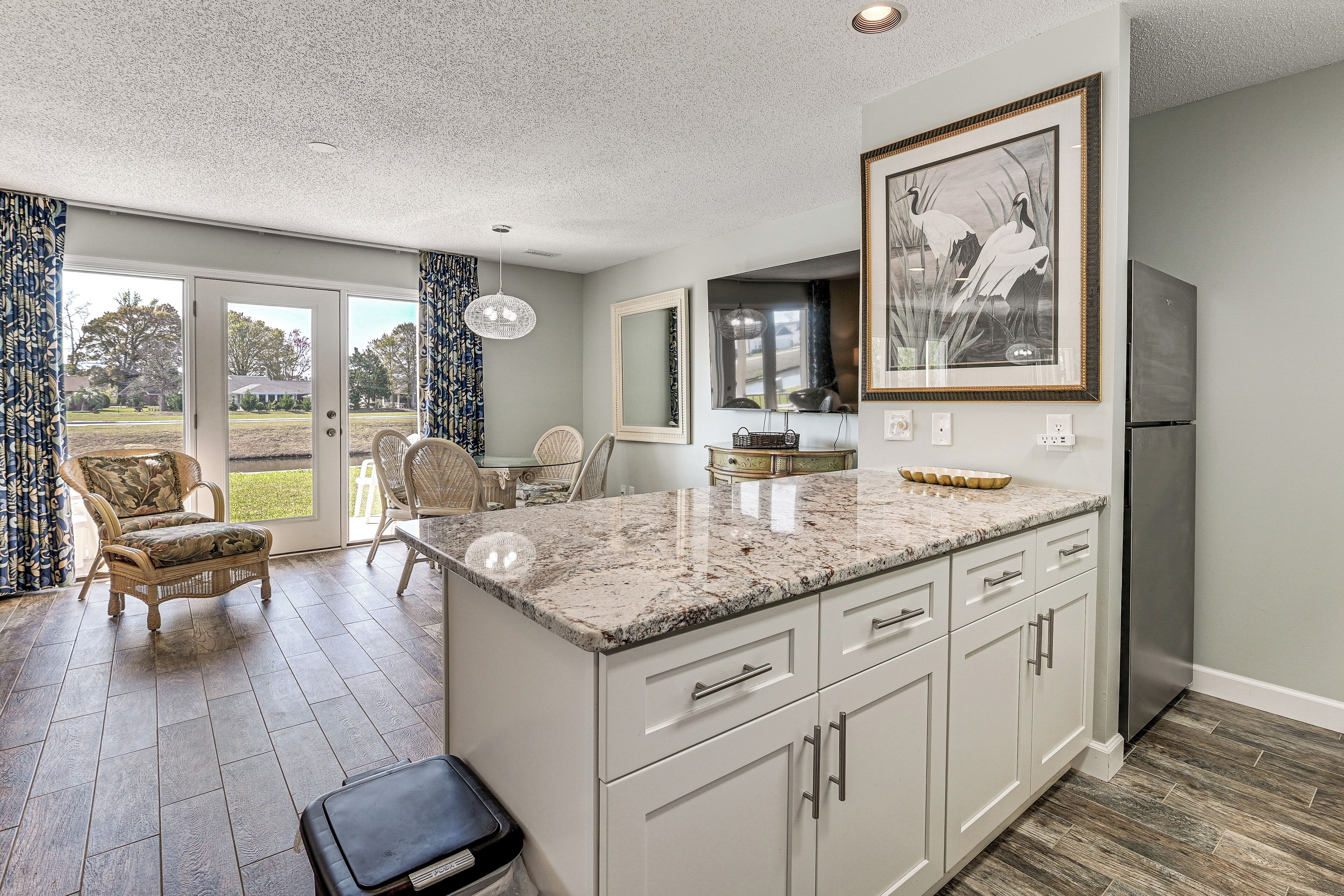 A kitchen with a marble countertop, white cabinets, and a stainless steel refrigerator. There is a dining area with wicker furniture and a view through glass doors.