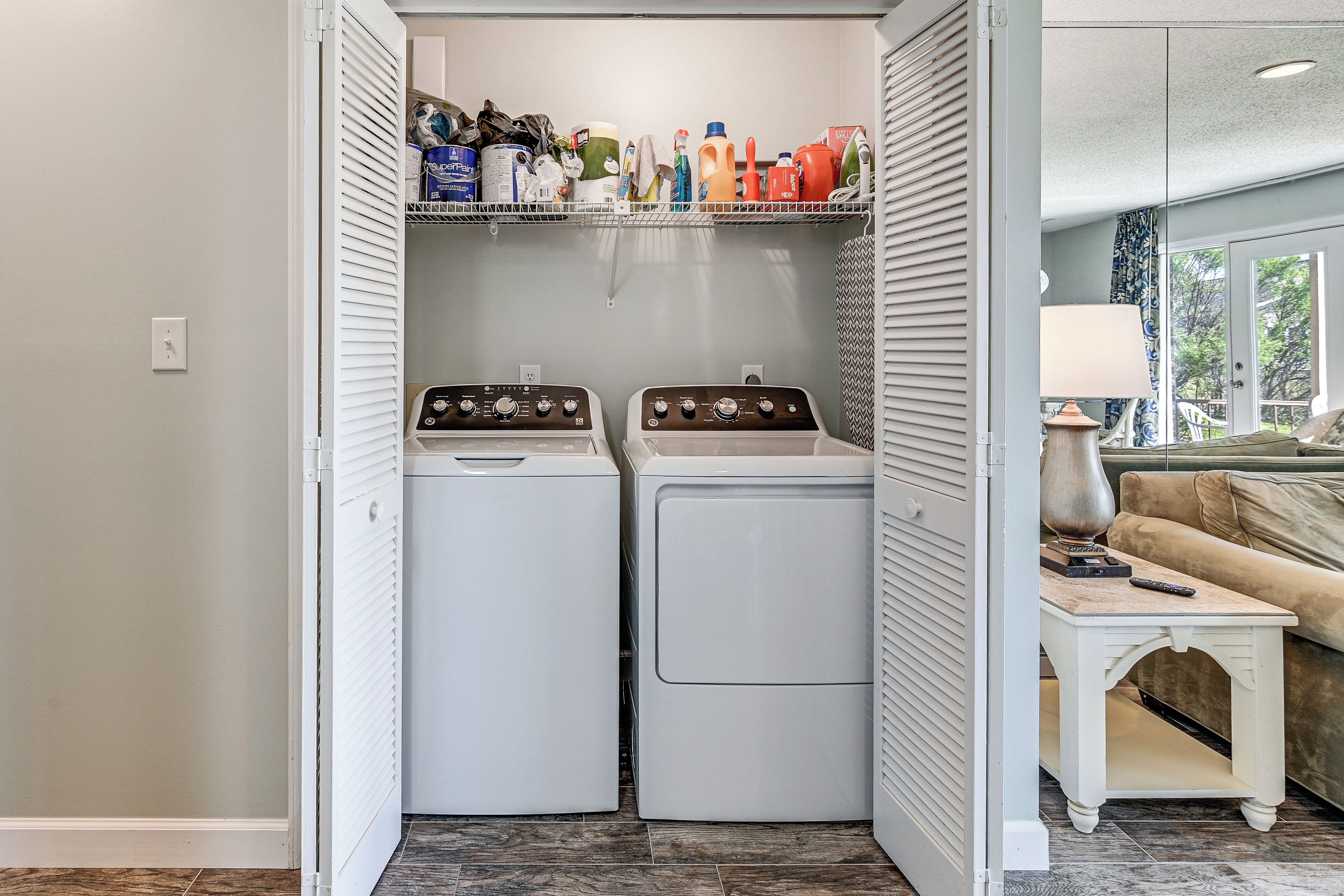 A laundry room with two front-loading washers and dryers, a shelf with cleaning supplies, and a small table with a lamp.