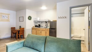 A living room with a green sofa, wooden dining table, and chairs, a kitchenette with a microwave, refrigerator, and cabinets, and a bathroom visible through an open door. - < 1 Mi to Boat Launch! Schroon Lake Basecamp (Schroon Lake)