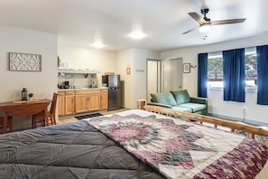 A bedroom with a bed, a desk, a kitchenette, and a view of the outdoors.