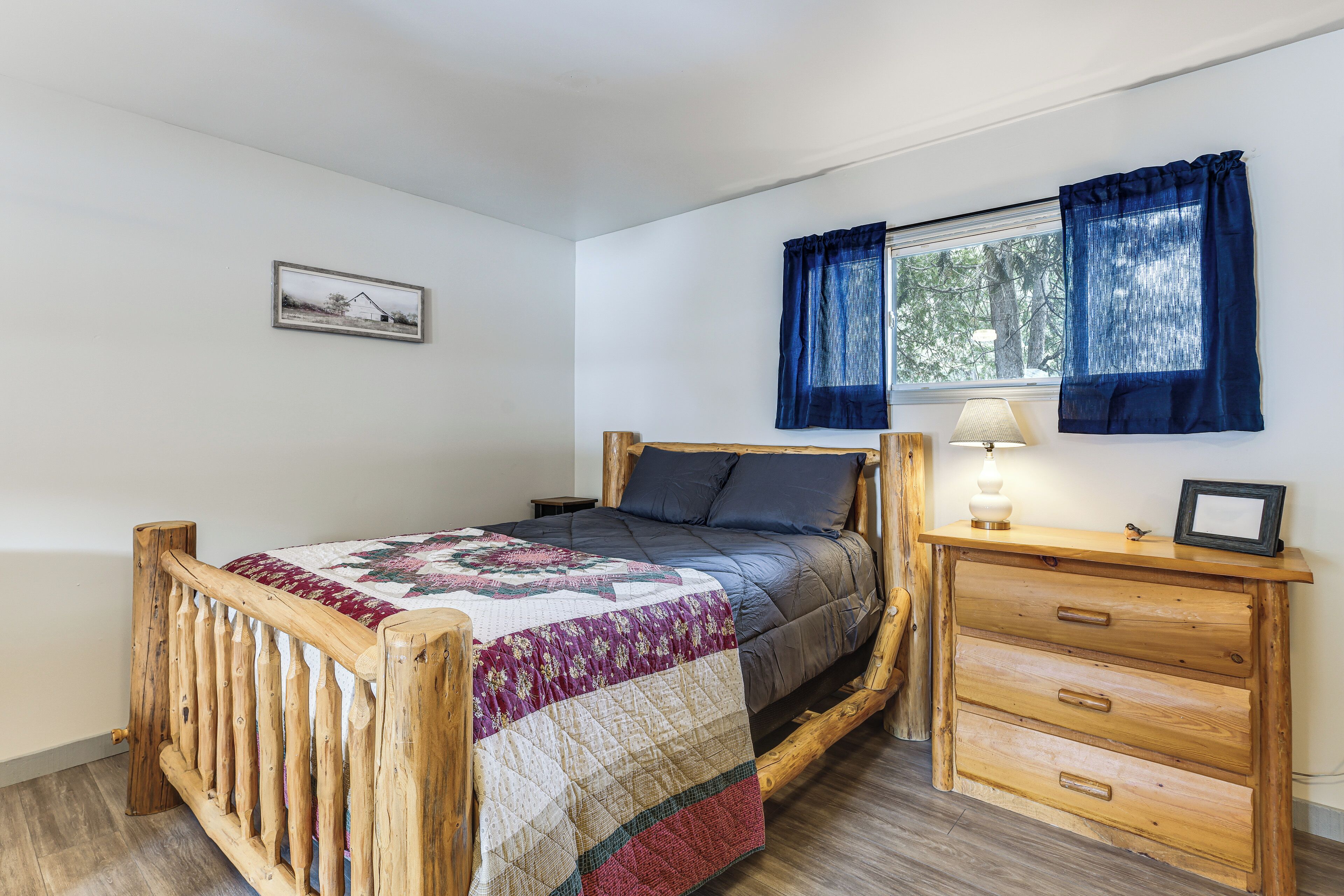 A wooden bed with a quilt, a bedside table with a lamp, and a window with curtains.