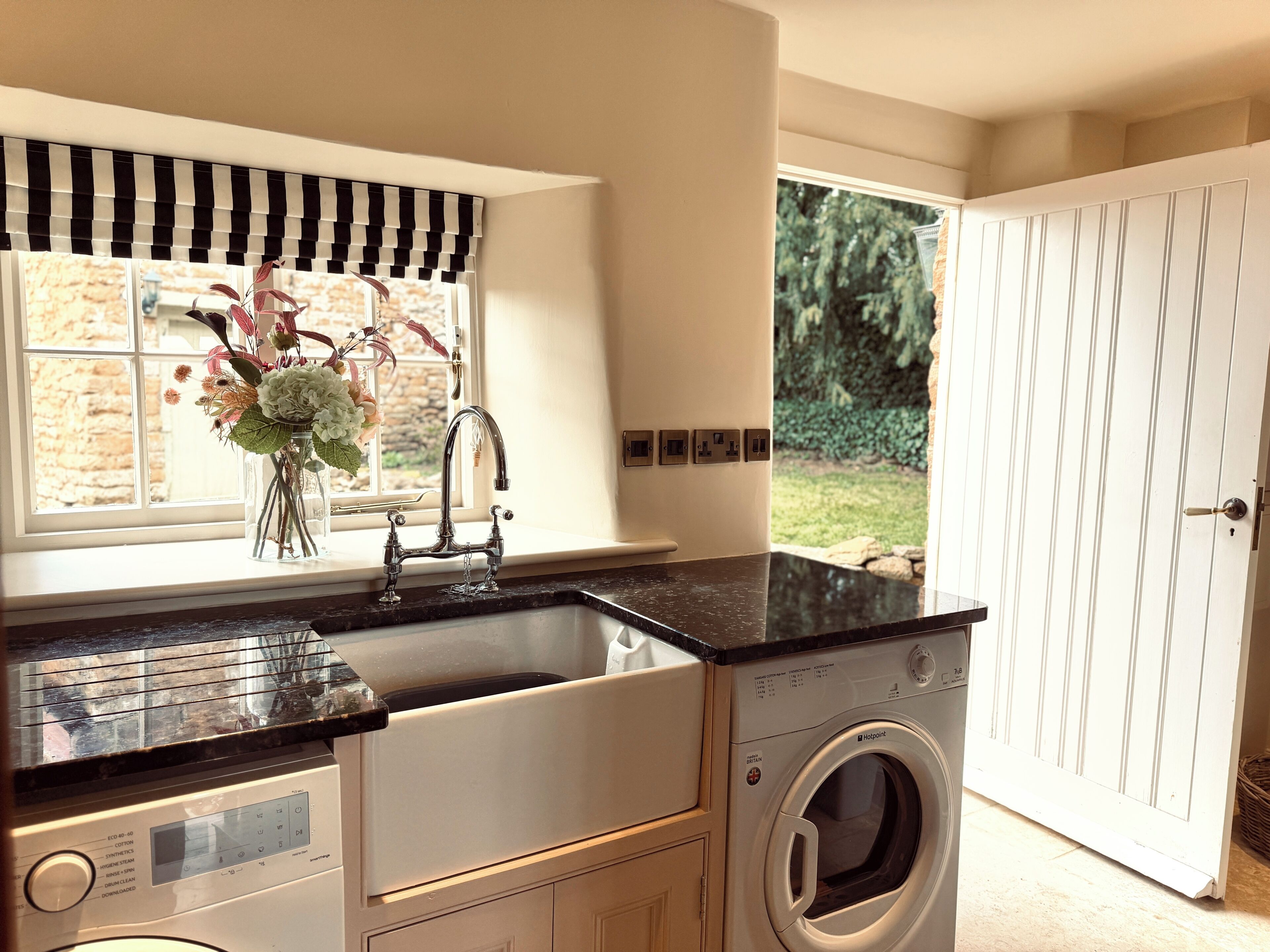 A laundry room with a washing machine, a sink with a black countertop, and a window with a black and white striped valance.