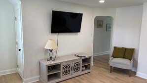 Un salon avec un téléviseur à écran plat fixé au mur, une console en bois sur laquelle est posée une lampe, et un fauteuil gris agrémenté d’un coussin vert.