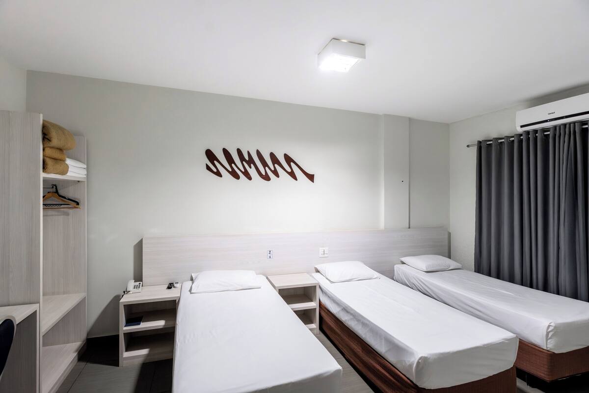 A hotel room with two single beds, a wardrobe, a bedside table with a lamp, and a wall-mounted air conditioning unit.