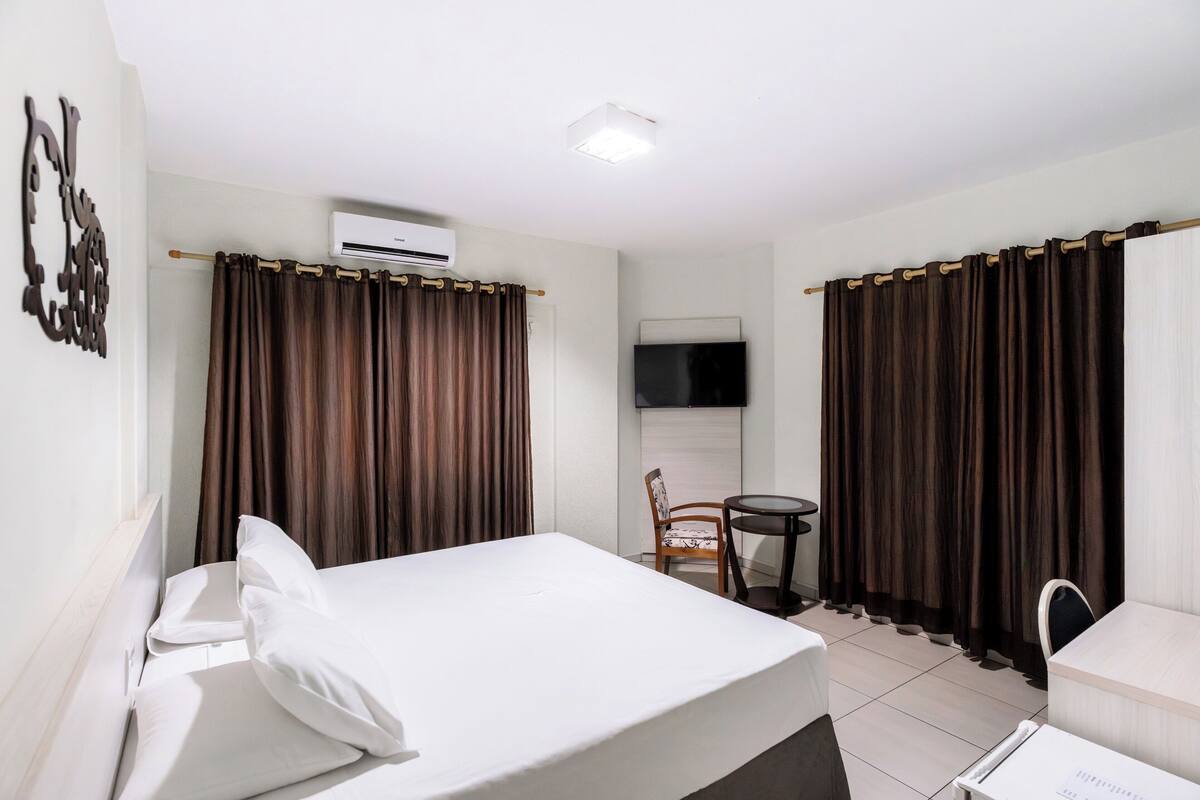 A hotel room with a bed, a chair, a small table, an air conditioning unit, a TV, and curtains.