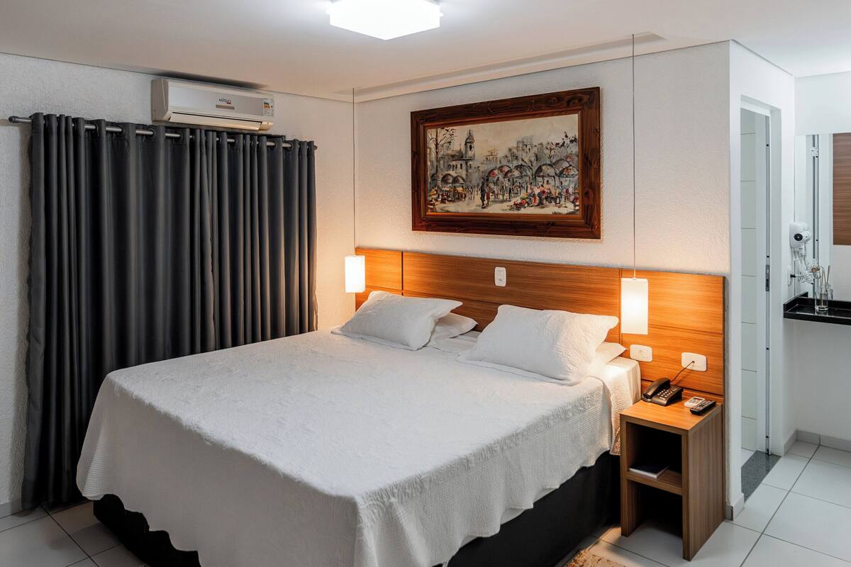 A hotel room with a bed, bedside tables, a painting, and an air conditioning unit.