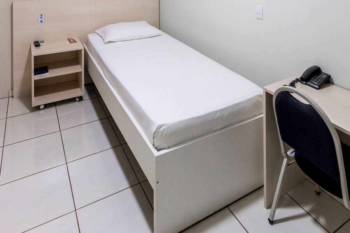 A single-bed room with a desk, a chair, a bedside table, and a ceiling light.