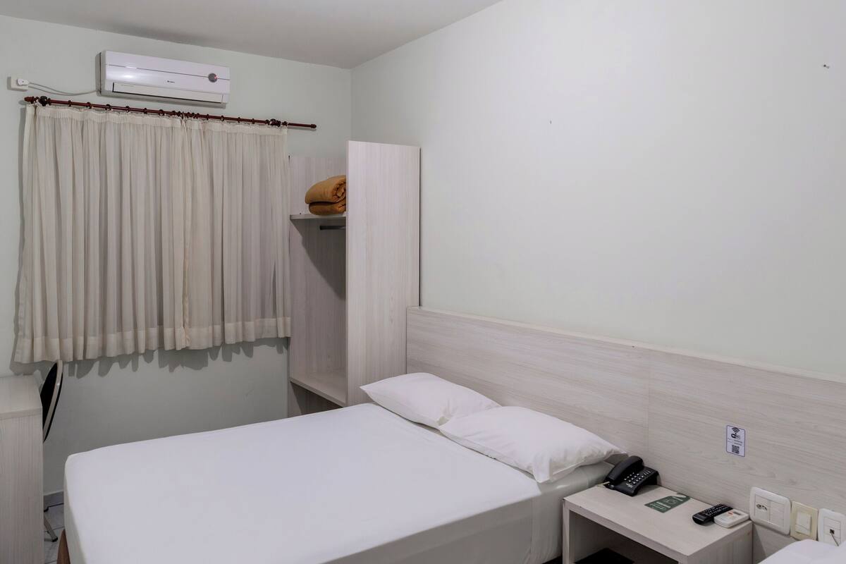 A hotel room with a single bed, a bedside table with a phone and remote, a wardrobe, and a curtain.
