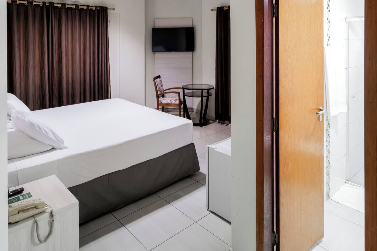 A hotel room with a bed, a desk, a chair, a TV, and an air conditioning unit.
