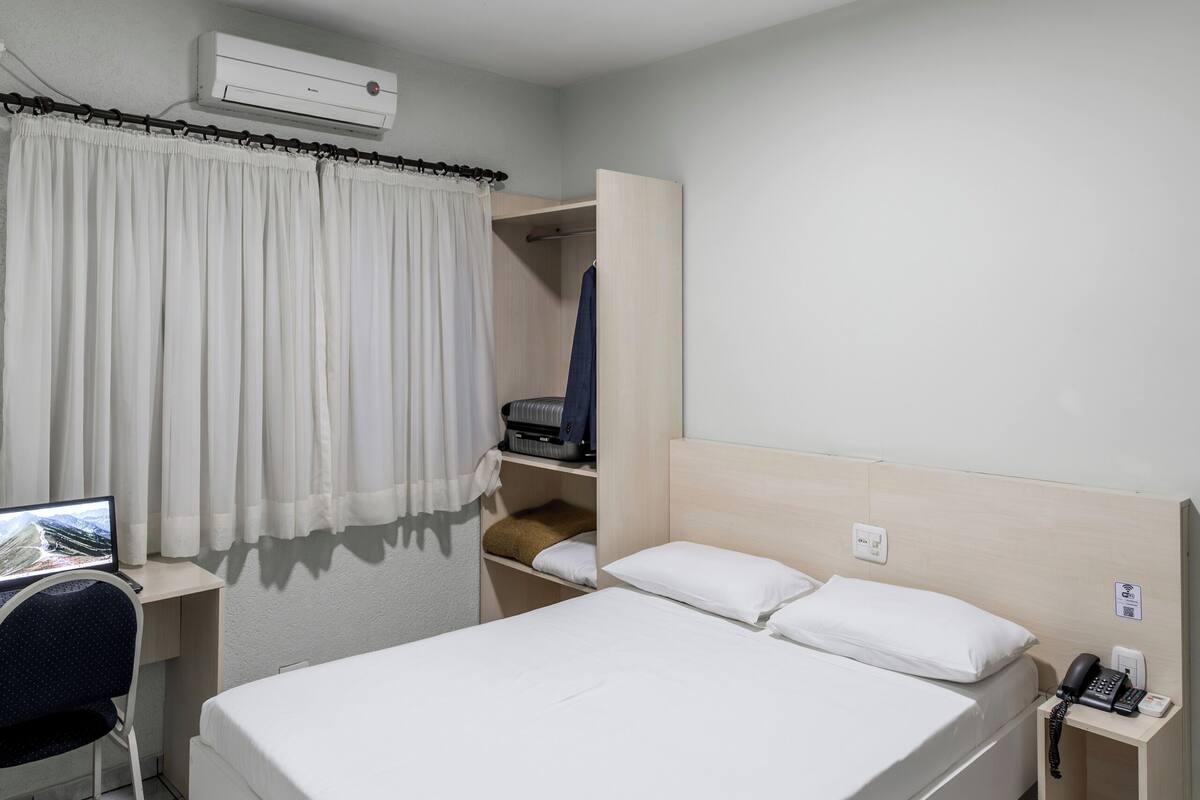 A hotel room with a bed, a desk with a chair, a wardrobe, a telephone, and an air conditioning unit.