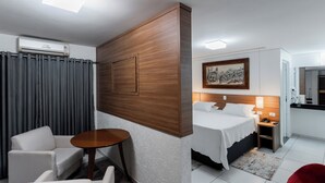 A hotel room with a bed, a desk, a chair, a small table, a painting on the wall, and an air conditioning unit.