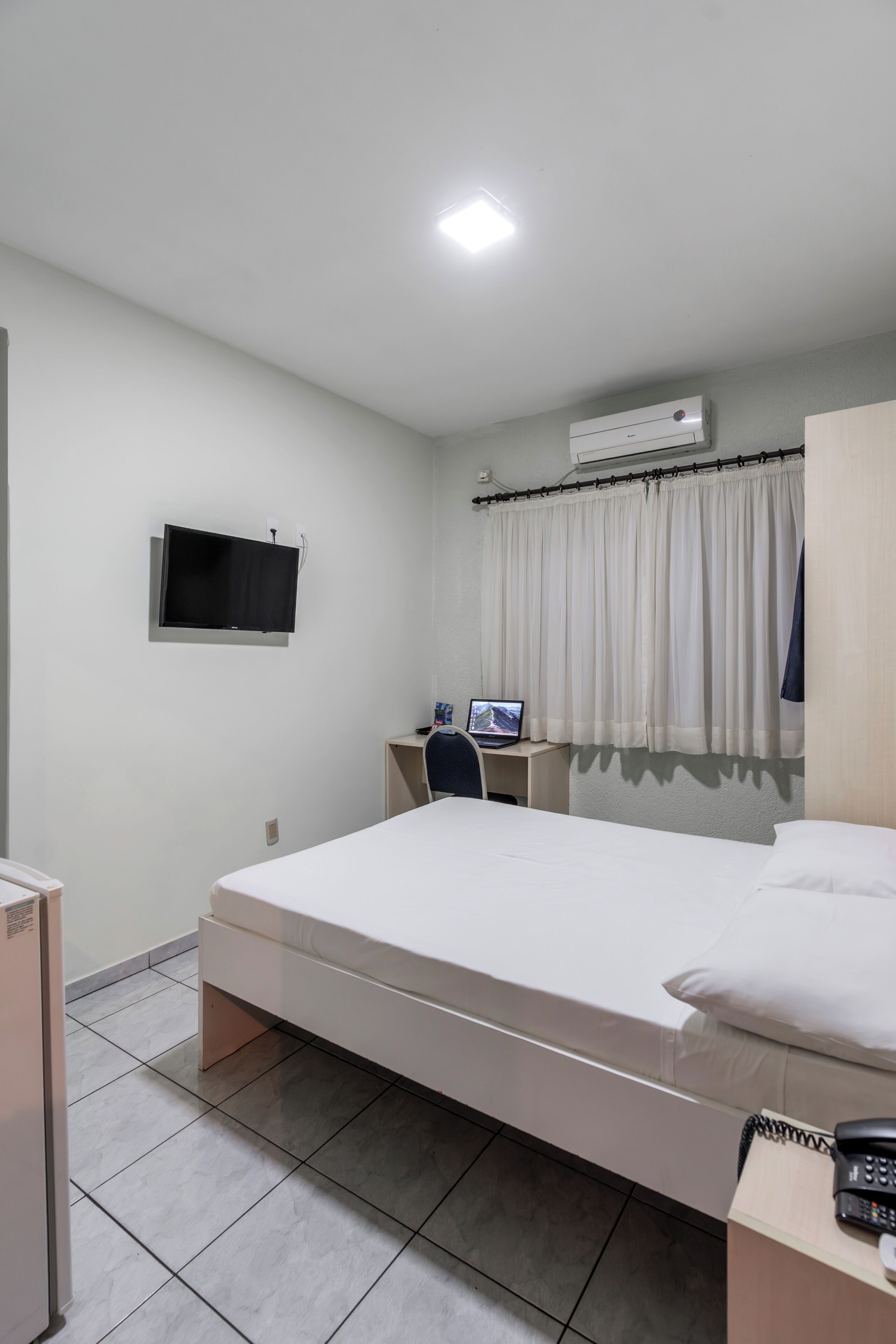 A hotel room with a bed, a desk with a computer, a TV, an air conditioner, and a telephone.