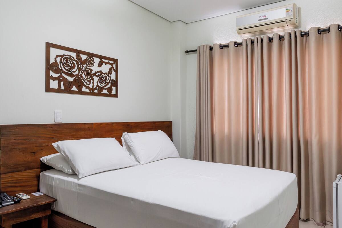 A hotel room with a bed, a nightstand, a wall-mounted air conditioner, and a window with curtains.