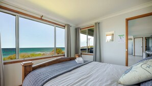 Un dormitorio con un ventanal grande con vistas al océano, una cama con colcha azul y blanca, y un cabecero de madera.