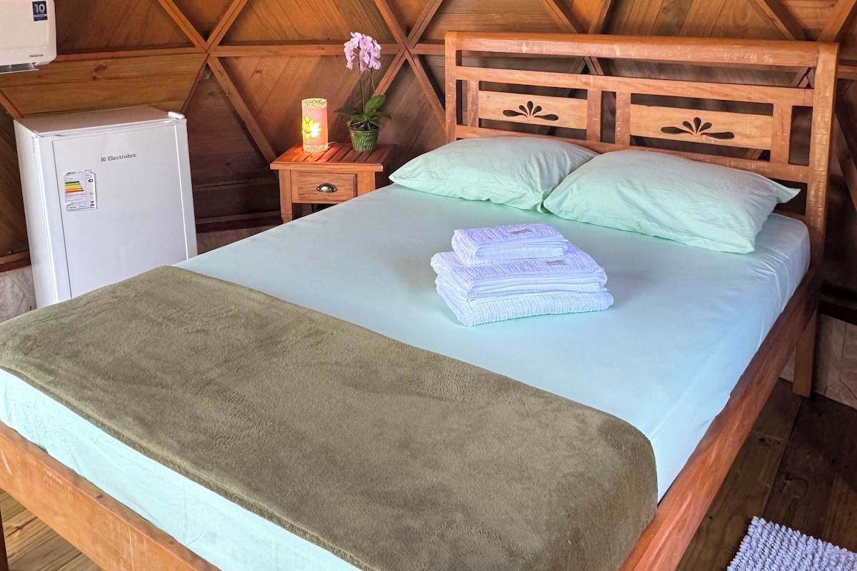 A bedroom with a wooden ceiling, a bed with green bedding, a nightstand with a lamp, and a small refrigerator.