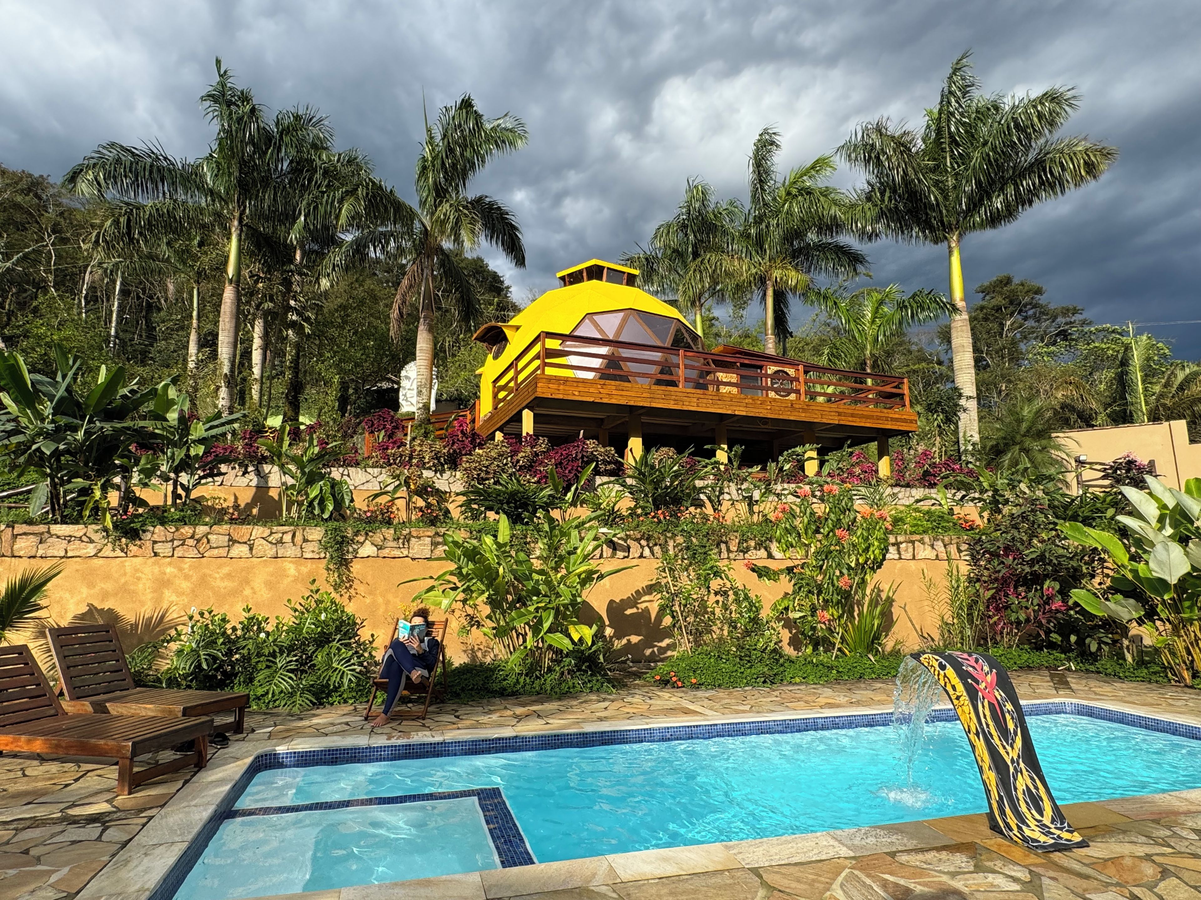 Área de piscina com um prédio amarelo, palmeiras e uma pessoa lendo um livro.
