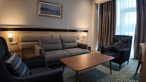 Une chambre d’hôtel avec un canapé, deux fauteuils, une table basse en bois, un tableau au mur et une fenêtre avec des rideaux.