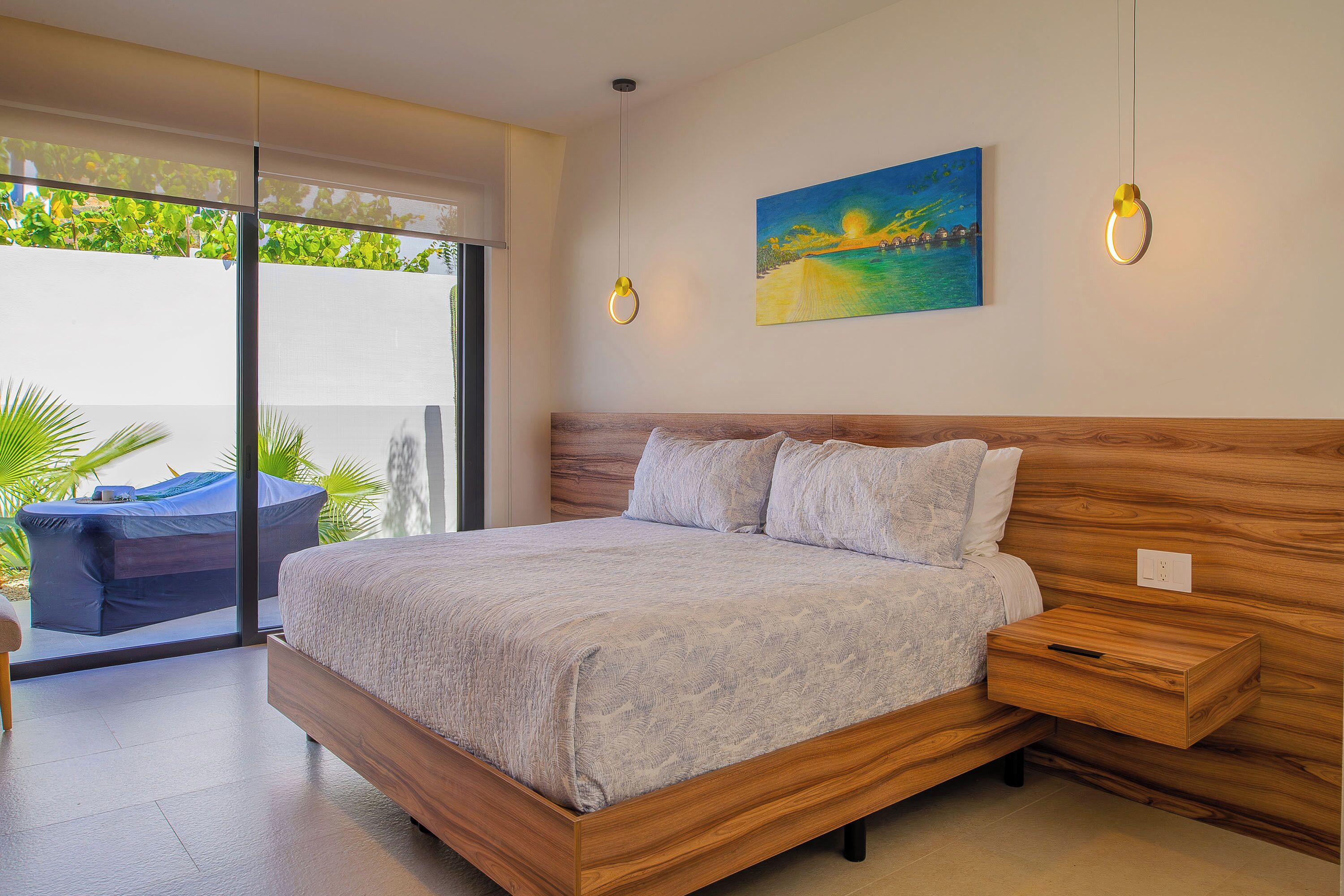 A bedroom with a large bed, a wooden headboard, a nightstand, a painting, and a view through a glass door.