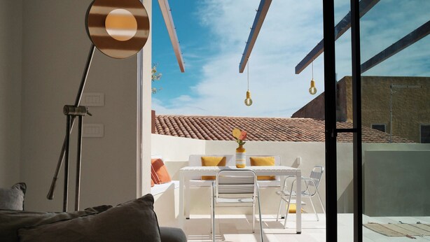 Ein modernes Wohnzimmer mit einer Couch, einem Esstisch mit Stühlen, einer Stehlampe und Blick auf ein Ziegeldach durch eine Glastür.
