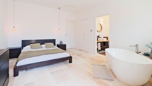 Ein modernes Schlafzimmer mit einem großen Bett, Nachttischen, einem Beistelltisch, einem begehbaren Kleiderschrank und einer freistehenden Badewanne.