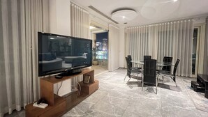 Een moderne woonkamer met een flatscreen televisie op een houten standaard, een eethoek met een glazen tafel en stoelen, en een stadsgezicht door grote ramen.