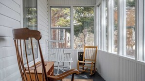 Ein Zimmer mit einem Holzstuhl, einem weißen Stuhl und einem Tisch. Große Fenster bieten Blick auf Bäume und Gebäude.