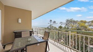 A balcony with a table and chairs, a white railing, and a view of trees and the ocean. - 2418 Windsor II - Oceanfront 4-Bed/4-Bath Villa - Fantastic Wrap Around Balcony! (Hilton Head Island)