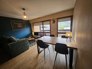 Un salon moderne comprenant un canapĂ©, une table Ă manger avec des chaises, un tĂ©lĂ©viseur et une grande fenĂȘtre avec des stores.