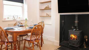 A dining area with a wooden table set for four, chairs, a fireplace with a wood-burning stove, and shelves with decorative items.